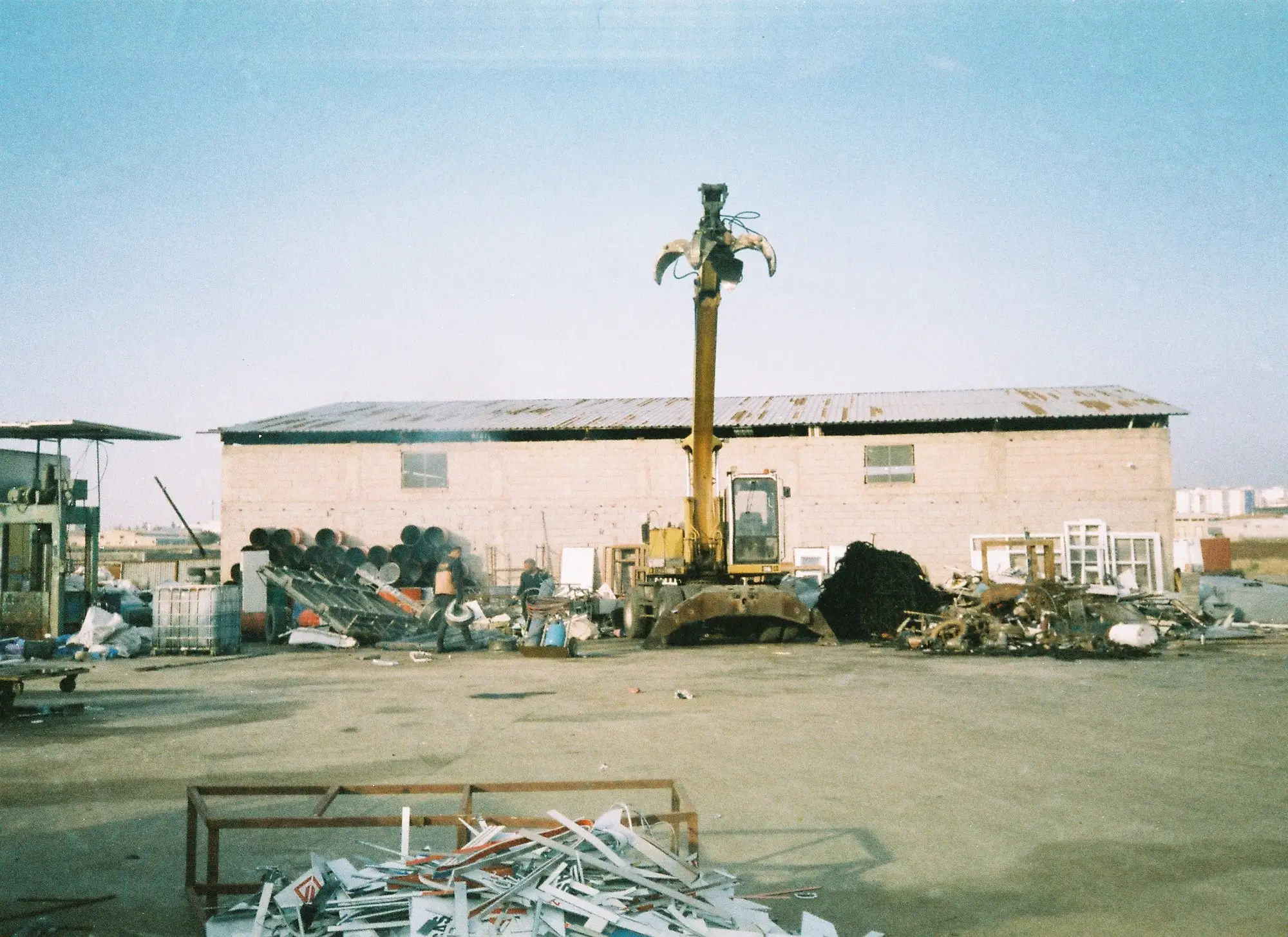 A material handler with a grapple standing on an open yard in front of a low shed, scrap piled to either side